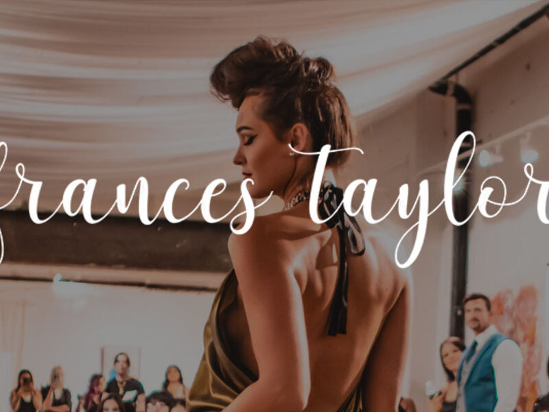 Woman in an elegant backless halter dress walking a runway at an indoor fashion show, with spectators and draped fabric ceiling in the background.