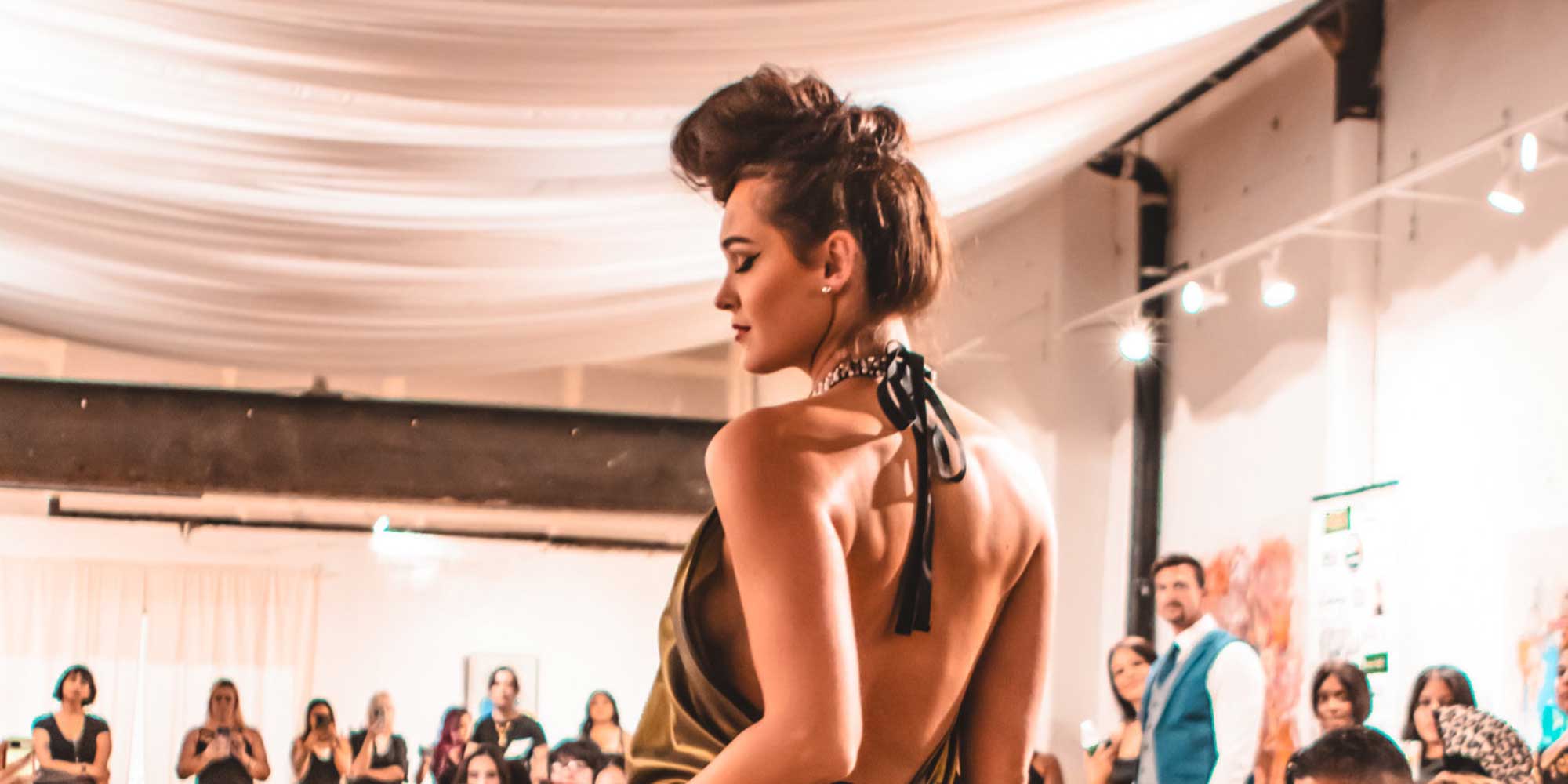 Woman in an elegant backless halter dress walking a runway at an indoor fashion show, with spectators and draped fabric ceiling in the background.