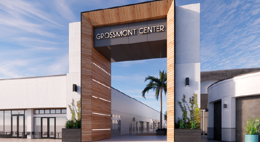 Modern shopping center entrance with a large wood-accented archway reading “Grossmont Center,” flanked by white buildings, planters, and a palm tree under a blue sky.